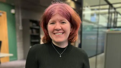 'We were sacked from Rockstar North for trying to unionise' Sarah Blackburn has shoulder-length pink and orange hair. She is smiling at the camera and wearing a black top and a silver necklace