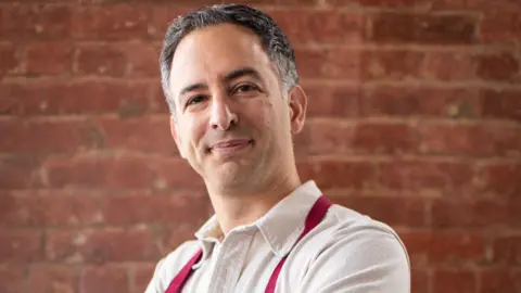 Ori Zohar Ori Zohar poses wearing a white shirt and red braces, standing in front of a brick wall.