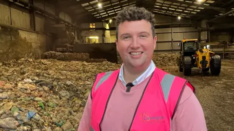 How many UK homes could be heated from cow manure? A man smiling in a pink jumper and fluorescent jacket that says 'Cannington Enterprises Ltd' on it. He is standing in a large shed which has what looks like bags of food waste in large piles behind him. There is also a yellow digger behind him.