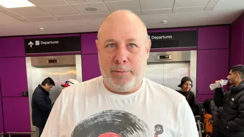 Flights resume at Edinburgh Airport after air traffic control outage Jeff is bald with a grey beard. He has blue eyes. He has a white t-shirt on. The airport is in the background.