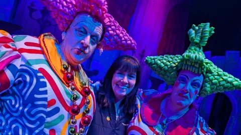 Pantomime is the bedrock of regional theatre, says Arts Council The owner and manager of The Academy Theatre in Birdwell stands between two panto dames who are wearing brightly coloured costumes and exaggerated make-up. The dame on the left is wearing a bright pink pointy plastic wig. The dame on the right is wearing green pointy plastic hair.