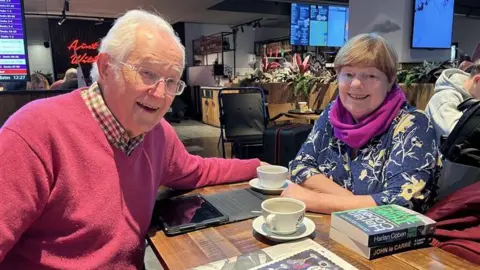 Flights resume at Edinburgh Airport after air traffic control outage Iver and Margot Morton sitting at a table with cups of coffee, books and a tablet on the table. Iver has grey hair and glasses and is wearing a dark pink jumper. Margot has short brown hair and is wearing a blue top with a floral pattern and a purple scarf.