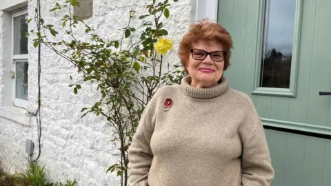 BBC Vanessa Corby is standing in front of her door. She is wearing a brown jumper with a poppy pinned to her right shoulder. She has short, styled reddish brown hair and black-rimmed glasses and smiles to the camera. Behind her is a white wall with a yellow rose bush growing up it. There is a green panelled door with a central window over her left shoulder.