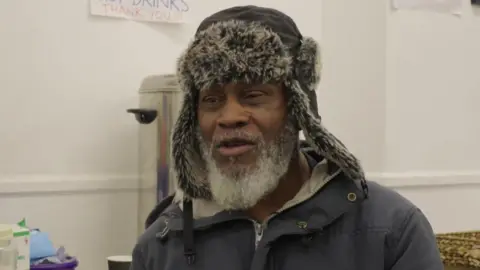 Brixton Soup Kitchen prepares for busy Christmas Elijah Augustus in a dark coat and hat with a fur trim. Behind him is a metal container for hot drinks.
