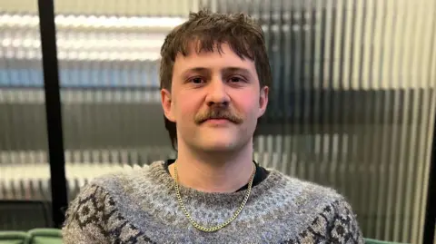 'We were sacked from Rockstar North for trying to unionise' Jordan Garland has short, brown hair and a moustache and is sitting in front of a distorted glass window. He is wearing a gold chain and a grey jumper with blue and black geometric prints