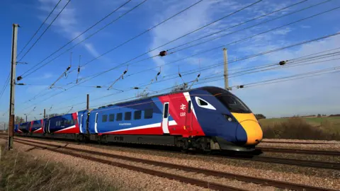 Department for Transport A train with the new red, white and blue branding - it is on a track somewhere in the countryside, with cabling overhead, a blue sky behind it, and fields stretching into the distance. 