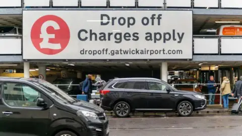 Getty Images A large sign at an airport car park saying Drop off charges apply with cars and people and cases below it