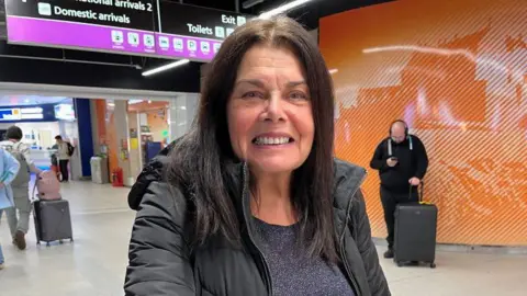 Flights resume at Edinburgh Airport after air traffic control outage Christine is smiling at the camera. She has long dark hair and is wearing a black coat and top. The airport is in the background.