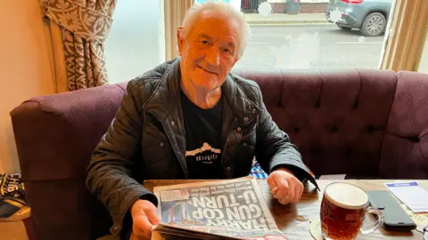 Pub regular Michael Tucker has swept-back white hair and is sitting at a table reading a paper with a pint of beer, and is looking up to the camera smiling. He is wearing a leather jacket and is sitting on a red sofa in front of the window.