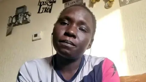 Martha has short cropped black afro hair and she is wearing a white, navy and pink top. She is sitting on a sofa and the wall behind her has various ornaments hanging on the wall.