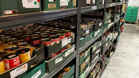 BBC A shelving unit in a warehouse. The shelves are full of cardboard crates which contain cans of food.