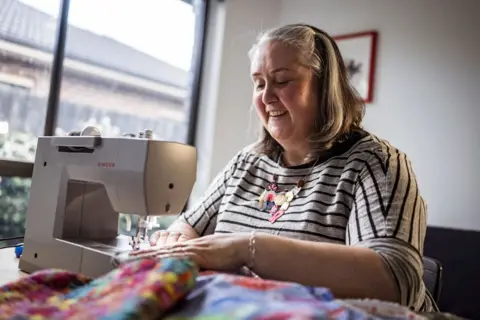 Chris Hopkins/BBC Maree O'Connor wearing a striped tshirt and colourful charm necklace, smiles as she is feeding fabric through a sewing machine.