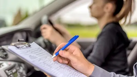 Getty Images A driving instructor holds a pen to a clipboard in a car whilst a young woman takes her test in the driving seat