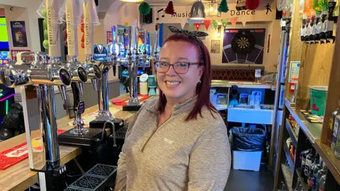 BBC Landlady Emma Holmes standing behind the bar smiling into the camera, photo is taken behind the bar with the taps to the left and spirits on shelves to the right.