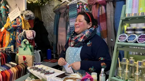 A woman wearing a Christmas tree headband, a colourful scarf and a white and blue apron and a very jolly face is behind the counter. In front of her is an array of tins of cream, and bottles with infusers in them. There is a large collection of leather purses and scarves to the side and behind her. 