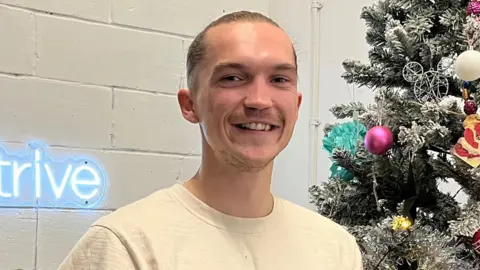 Connor Bennett/BBC Lewis Wardale, a man who is looking directly at the camera and smiling. He is standing in front of a white painted brick wall with a decorate Christmas tree on his right. 