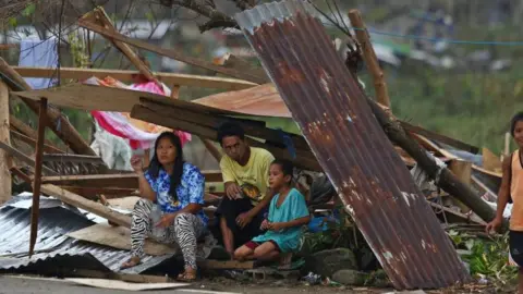 Getty Images A family of three crouches for shelter under a rusted piece of galvanised roof, by a roadside in the Philippines in the wake of Typhoon Rai in December 2021
