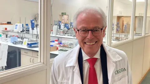 BBC A man wearing a suit, red tie and white lab coat, with the word Croda written on it in green letters, stands in a corridor outside a lab, which can be seen through large windows. Rows of desks contain various chemical equiment.