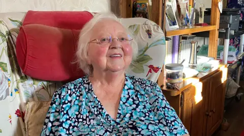 Anne Hutchinson is a woman in her 80s sitting in an armchair. She is smiling and is wearing a blue top and has white hair and glasses with pink cheeks. Behind her is a dresser with books and photographs on display.