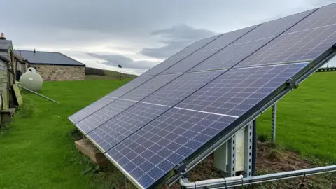 A frame on the ground holds eight solar panels in two rows of four in the foreground of the picture. It sits on a huge expanses of grass and in the distance is a wing of the stone cottage. The circular end of a green LPG gas tank is also visible, between a shed and the main building.
