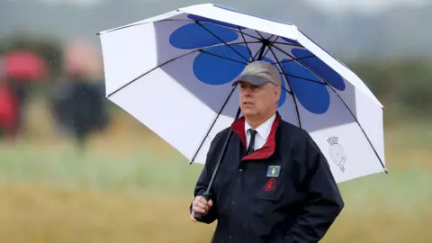 Getty Images Andrew Mountbatten-Windsor follows the action at the Open Championship while taking shelter under a blue and white golf umbrella. He is wearing a blue waterproof jacket and navy pin name badge pinned to his left breast. He is also wearing a grey baseball cap.