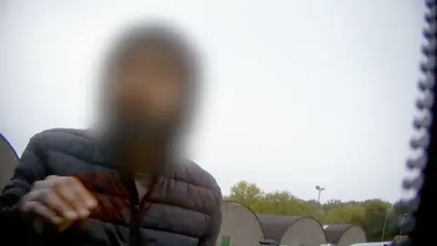 'Throw the parcel at the back door' Man with a blurred face wearing a black anorak. There is grey sky behind, with trees in the distance. The image was taken undercover, and part of a zip is visible on the right hand side.