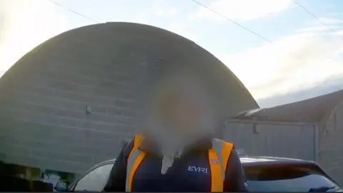 'Throw the parcel at the back door' A woman with a blurred face wearing an orange high-vis gilet and a blue fleece. There is the arc of the roof of a building in the background.