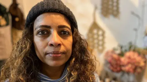Shiromi wearing a dark knitted hat and a blue-and-white striped long-sleeve top. She has long brown wavy hair. The blurred background shows shelves with bottles, decorative items, and an arrangement of pink flowers.