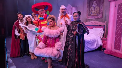 Pantomime is the bedrock of regional theatre, says Arts Council The cast of Beauty and the Beast are stood on the stage of the Lawrence Batley Theatre in Huddersfield. The dame, Betty Bon Bon, is stood centre stage in a brightly coloured costume which looks like a jar of jelly beans. The other cast members include Belle who is dressed in a white nightie, her sister in a bright pink glittery dress with pink fur trim - and the baddie who is a witch in a black and red dress.