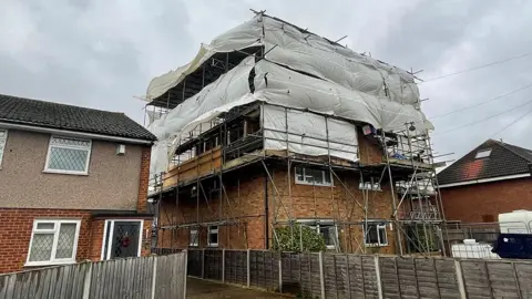 Full view of the block of flats from ground level. The building is covered with scaffolding and big sheets of plastic rise over the first-floor windows. There is a fence in front of the property - and houses either side.