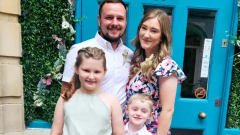 Laura Winter A picture of Tom, Laura, Amelie and Evan Winter - all smiling as they stand in front of a blue door with a floral display on either side of the door.