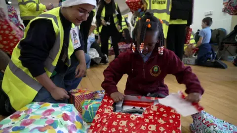 A young girl opens a wrapped Christmas present on the floor of a school hall while a volunteer in a high-visibility vest watches, with other children, volunteers and gifts visible in the background.