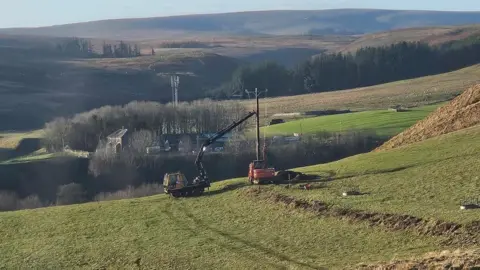 Shona Anderson A cherry picker is inserting an electricity pole into the ground in a field. In the distance is a cluster of farm buildings and beyond that open moorland 