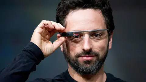 Bloomberg via Getty Images Sergey Brin wears a black long-sleeved top and Google Glass. He is looking directly into the lens of the camera capturing him, with one hand hovering over the right side of Google's digital glasses. On the right lens of the glasses, beside a black box containing the glasses camera, is a small red and black digital display.