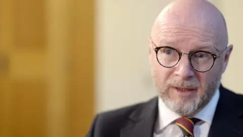 'Throw the parcel at the back door' Liam Byrne has a greyish beard and wears a suit and glasses. He sits to the right of the camera and is photographed as he is being interviewed.