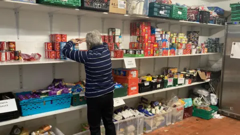 Empty shelves fill Coventry food hub volunteers with dread A volunteer is stacking shelves in the food hub store room which shows lots of other food items on the shelves and in crates.