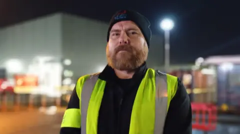 Workers go on strike at Guinness canning factory Billy looking at the camera. He has a beard and is wearing a beanie hat and high visibility vest over a black jacket. He is standing outside.
