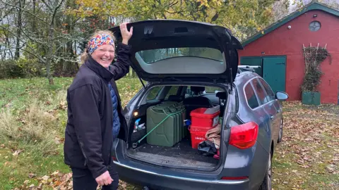 Doris Englemayer is standing at the rear or her car. Inside the boot you can see three jerrycans of petrol. She is in her 50s, wearing a headband, a jacket and a dark coat and is smiling.
