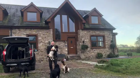 Malcolm Hogg Malcolm Hogg is standing outside his house with a spaniel in his arms and another one at his feet. Next to him is a car with an open boot. The house is new but built in a traditional style. He is a man in his sixties wearing black trousers and top.