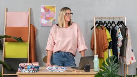 Nerida Hansen Nerida Hansen, wearing a pink top and blue jeans, stands next to a desk with a laptop open on it. She is holding a fabric sample print on a piece of paper in one hand. There are four printed fabrics piled on the side of the desk. A rack with clothes on hangers stands behind her. 