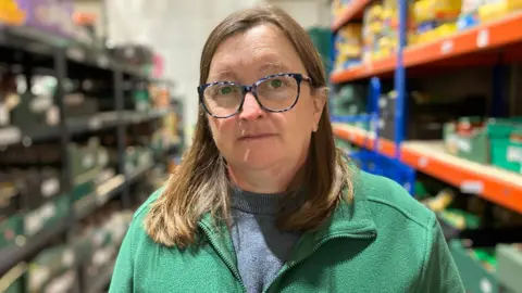A woman with long brown hair and glasses, wearing a green fleece and blue jumper, stands in between two shelves containing crates of food.