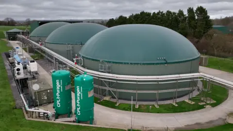 How many UK homes could be heated from cow manure? A biogas plant surrounded by grass. It is a large green structure made up of three domes. There are two green cylinders at the front of the image, saying CPL Puragen on them. There are trees in the background.
