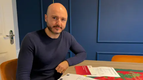 BBC Sevak Maljian sat at a table with various papers in front of him. He is bald with a dark beard, wearing a navy jumper.