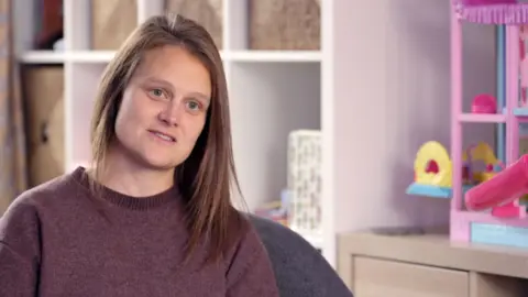 'Throw the parcel at the back door' Becky has long brown hair and wears a dark purple top. Behind her is a Barbie doll house and a large white shelving unit.