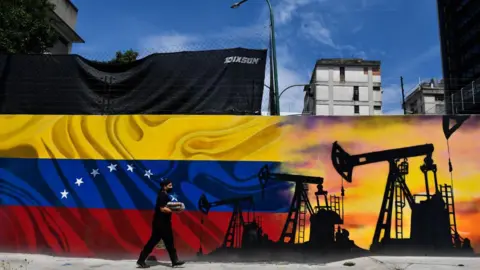 AFP via Getty Images A man wearing a face masks walks past a mural depicting an oil pump and the Venezuelan flag in a street of Caracas, on May 26, 2022. (Photo by Federico PARRA / AFP)