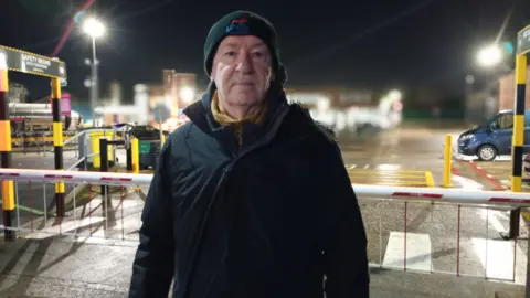 Workers go on strike at Guinness canning factory Michael looking at the camera. He is standing outside wearing a black coat and a beanie hat with unite embroidered on it.