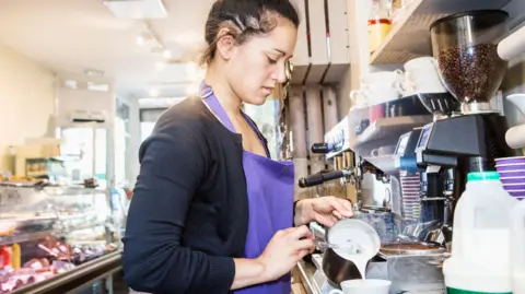 Getty Images A woman wearing a purple apron works in a cafe. She is pouring milk into a mug next to a coffee machine