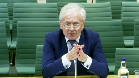 UK Parliament Prof David Miles sitting down giving evidence to MPs. He is wearing a blue suit, white shirt and grey tie. He has white hair and is wearing glasses. He is speaking while placing his hands together.