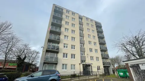 LDRS A tower block, with a blue car parked in front of it. There is an Asda supermarket behind the building, to the left of the image, and an outbuilding and a set of green industrial bins on the right hand side of the image.