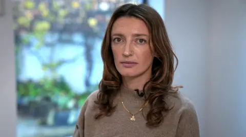 BBC Claire Corkery, a woman with long hair, wearing a beige jumper and a gold necklace, standing in her dining room.